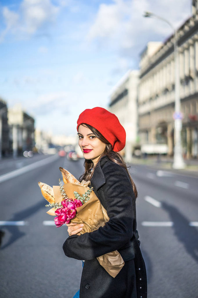 Young beautiful girl Frenchwoman brunette in a red beret and a black coat goes along the street of the European city with a bouquet of flowers and French baguettes. A happy smile and a charming look, the French style of Paris - Fotó, kép