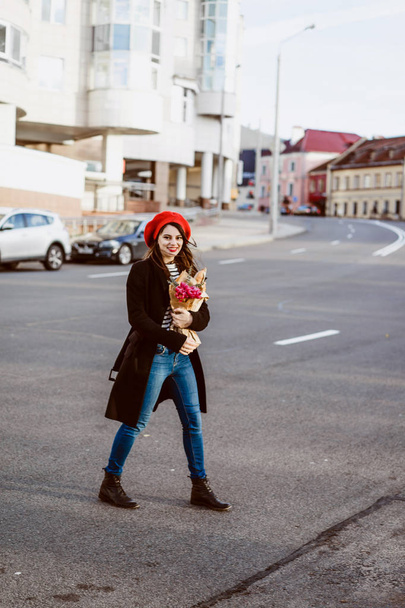 Young beautiful girl Frenchwoman brunette in a red beret and a black coat goes along the street of the European city with a bouquet of flowers and French baguettes. A happy smile and a charming look, the French style of Paris - Фото, изображение