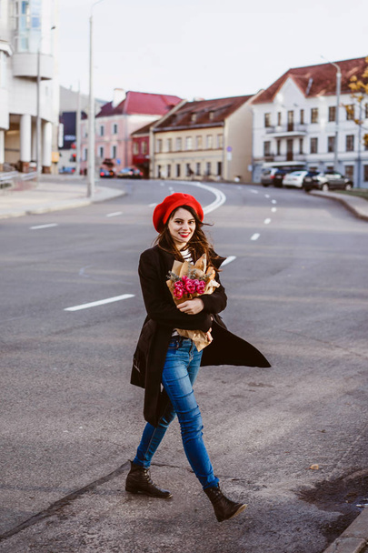 Young beautiful girl Frenchwoman brunette in a red beret and a black coat goes along the street of the European city with a bouquet of flowers and French baguettes. A happy smile and a charming look, the French style of Paris - Foto, immagini