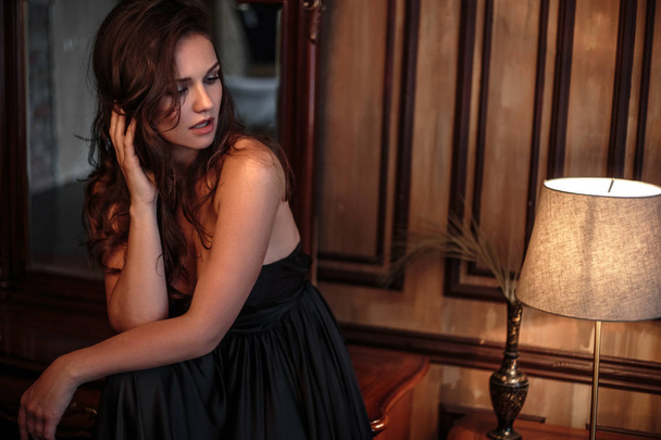 Portrait of young woman in elegant silk black dress posing in a dark interior, fashion beauty photo - Zdjęcie, obraz