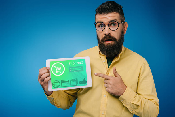 shocked bearded man pointing at digital tablet with shopping app, isolated on blue - Photo, Image