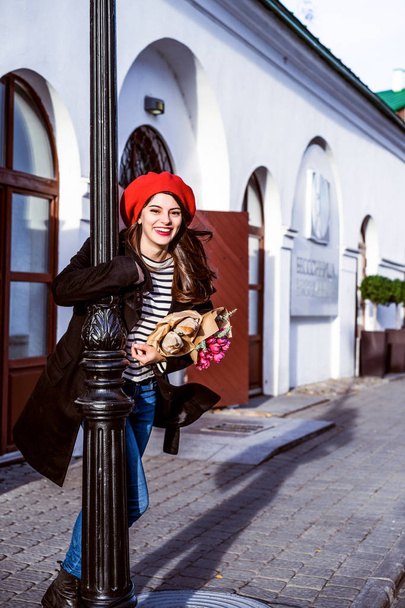 Genç güzel kız Fransız kadın esmer kırmızı bere ve siyah bir ceket Avrupa şehri Street'te bir buket çiçek ve Fransız baget ile gider. Mutlu bir gülümseme ve büyüleyici bir bak, Paris Fransız tarzı - Fotoğraf, Görsel