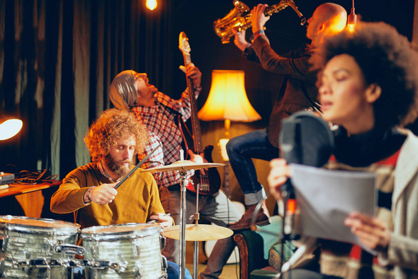 Mixed race woman singing. In background band playing instruments. Home studio interior. Selective focus on background. - Photo, Image