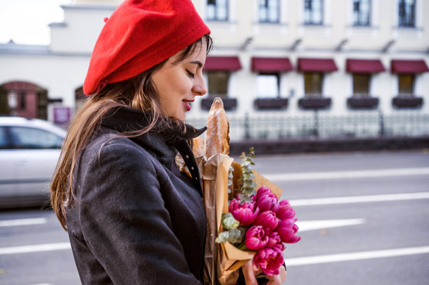 Young beautiful girl Frenchwoman brunette in a red beret and a black coat goes along the street of the European city with a bouquet of flowers and French baguettes. A happy smile and a charming look, the French style of Paris - 写真・画像