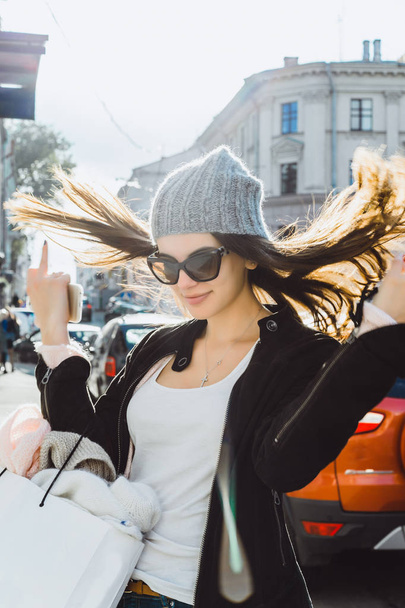 a beautiful brunette girl in sunglasses, a knitted cap, and a charming cute smile walks the streets of a European city, shopping, a big white cardboard package, hair develops, a smartphone in her hand - Фото, зображення