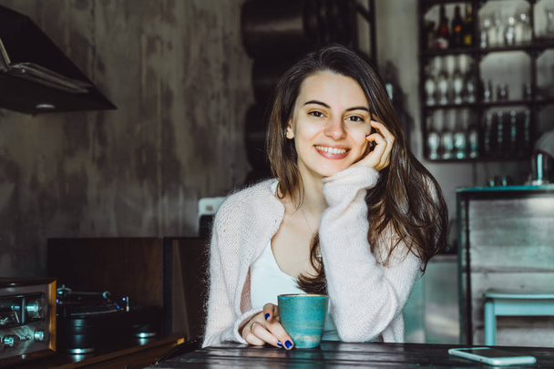 a beautiful brunette girl with a charming smile and expressive brown eyes drinking coffee and using a smartphone - Zdjęcie, obraz