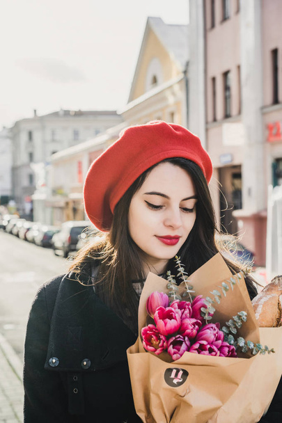 Young beautiful girl Frenchwoman brunette in a red beret and a black coat goes along the street of the European city with a bouquet of flowers and French baguettes. A happy smile and a charming look, the French style of Paris - Foto, Imagen