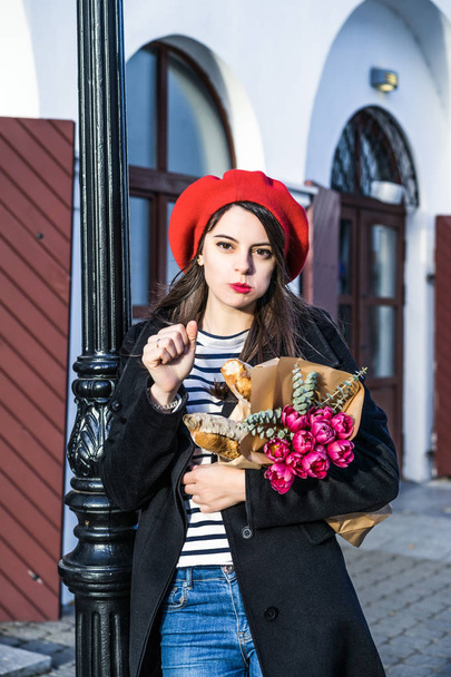 Young beautiful girl Frenchwoman brunette in a red beret and a black coat goes along the street of the European city with a bouquet of flowers and French baguettes. A happy smile and a charming look, the French style of Paris - Photo, image