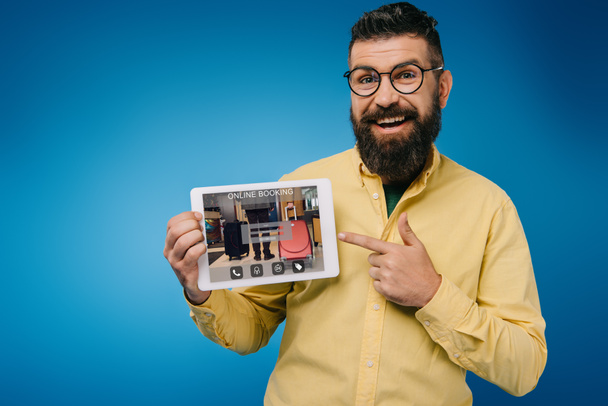 счастливый бородатый мужчина, указывающий на цифровую табличку с приложением онлайн-бронирования, изолированный на голубом фоне
 - Фото, изображение