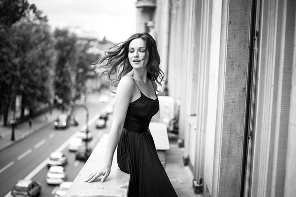 Black white portrait of young woman in elegant silk black dress lookingto a city from a balcony, fashion beauty photo - Фото, изображение