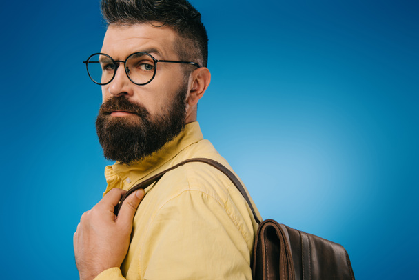 serious bearded man in eyeglasses with backpack, isolated on blue - 写真・画像