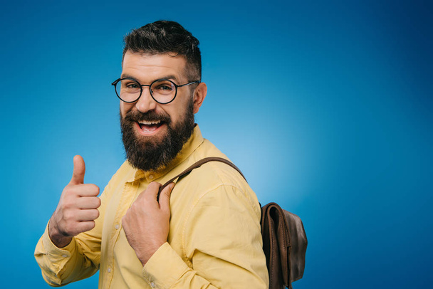cheerful bearded man with backpack showing thumb up, isolated on blue - Foto, Imagen