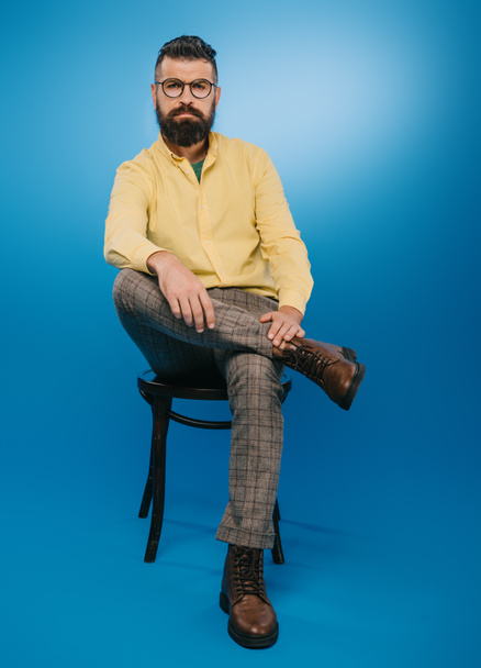 стильный бородатый мужчина в очках, сидящий на стуле изолированный на синий
 - Фото, изображение