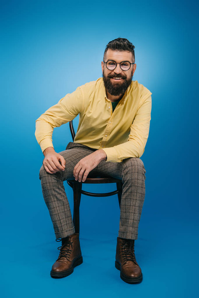 smiling bearded man in eyeglasses sitting on chair isolated on blue - Foto, imagen