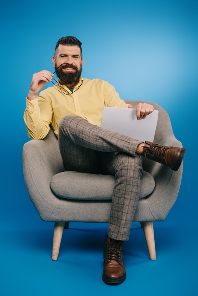 handsome smiling businessman sitting in armchair with laptop on blue - Fotografie, Obrázek