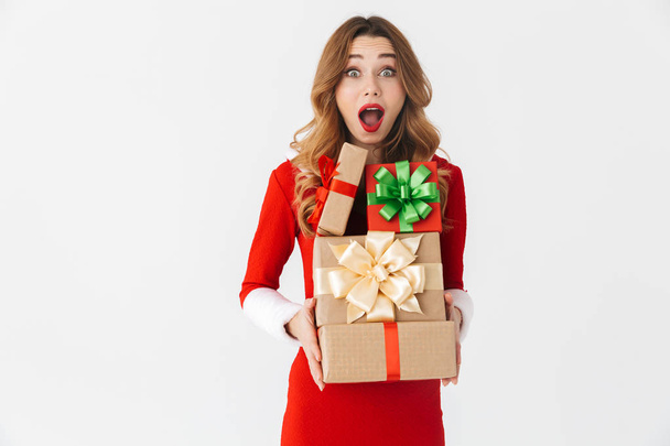 Excited young girl wearing Christmas dress standing isolated over white background, holding stack of present boxes - Foto, afbeelding