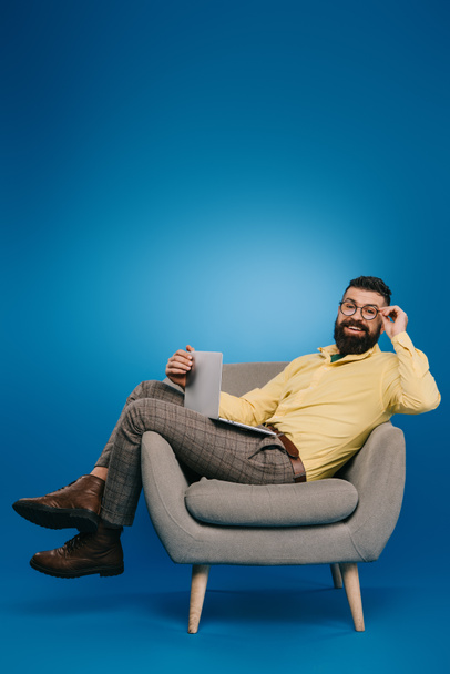 smiling bearded businessman sitting in armchair with laptop on blue - Zdjęcie, obraz