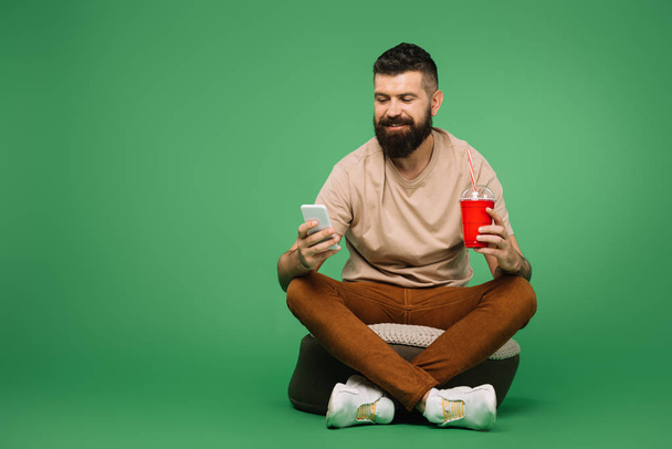 homme barbu souriant avec soda en utilisant un smartphone, isolé sur vert
 - Photo, image