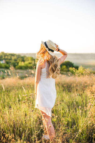 Lovely young girl with blond hair in a white sundress and a straw hat in the summer in a field at sunset on a sunny day - Foto, afbeelding