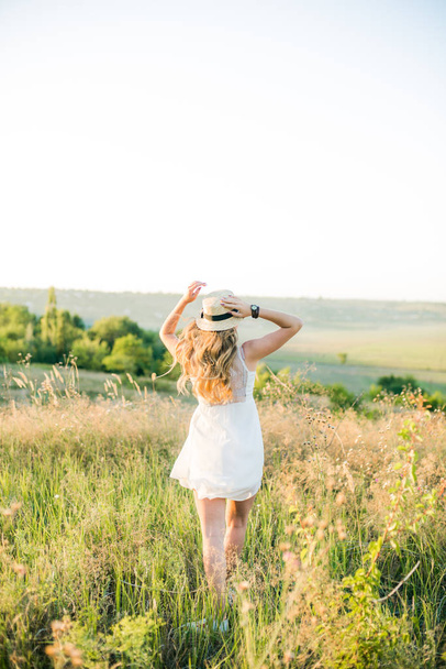 Lovely young girl with blond hair in a white sundress and a straw hat in the summer in a field at sunset on a sunny day - Foto, afbeelding