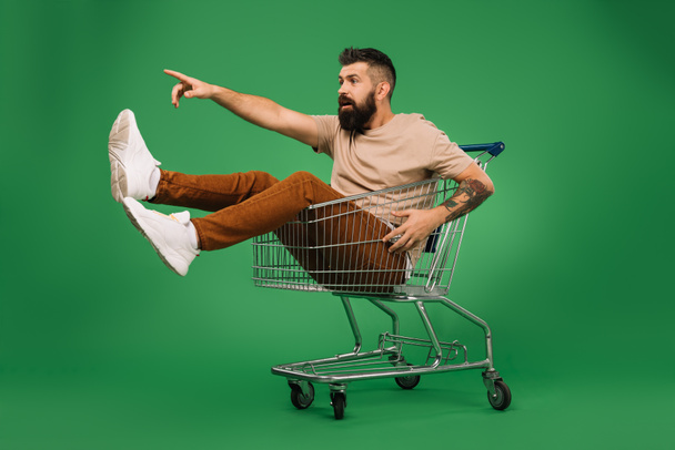 vzrušený pohledný muž ukazuje něco, zatímco sedí v nákupní košík izolované na zelené - Fotografie, Obrázek