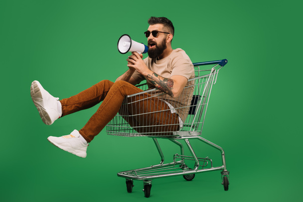 bearded man with megaphone sitting in shopping cart isolated on green - Φωτογραφία, εικόνα