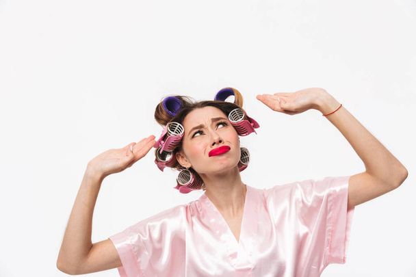 Upset housewife with curlers in hair wearing robe standing isolated over white background, looking up - Fotó, kép