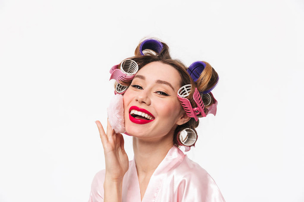 Beautiful housewife with curlers in hair wearing robe standing isolated over white background, using powder puff - Valokuva, kuva
