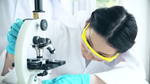 Group of chemists working in a lab. Young asian female chemists with senior caucasian chemist working together in lab, looking into microscope. Science concept. - Filmati, video