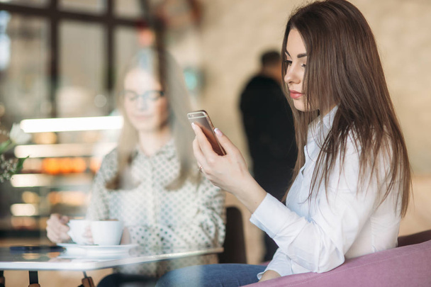 Две девушки сидят в кафе, пьют капучино и пользуются смартфоном. Современная девушка
 - Фото, изображение