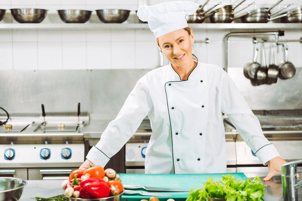 hermosa mujer chef en uniforme sonriendo y mirando a la cámara durante la cocina en la cocina del restaurante
 - Foto, Imagen