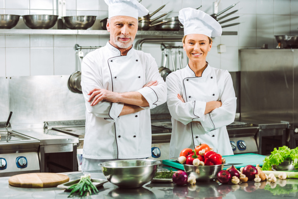 mannelijke en vrouwelijke chef-koks in uniform met armen gekruist tijdens het koken in de keuken van het restaurant - Foto, afbeelding