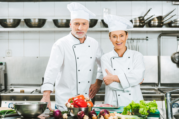 θηλυκά και αρσενικά σεφ με τη στολή με τα χέρια σταυρωμένα βλέπουν φωτογραφική μηχανή κατά τη διάρκεια της μαγειρικής σε κουζίνα εστιατορίου - Φωτογραφία, εικόνα