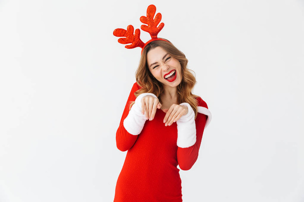 Portrait of flirting woman 20s wearing Santa Claus red costume and deer ears smiling and standing isolated over white background - Photo, Image