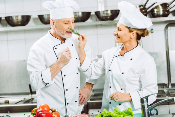 lächelnde Köchin mit Rosmarinkraut in der Nähe eines Mannes beim Kochen in der Restaurantküche - Foto, Bild