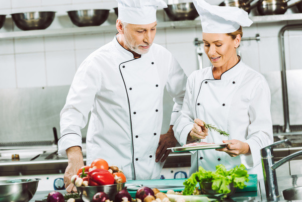 female and male chefs in double-breasted jackets and hats decorating dish with rosemary while cooking in restaurant kitchen - Photo, Image
