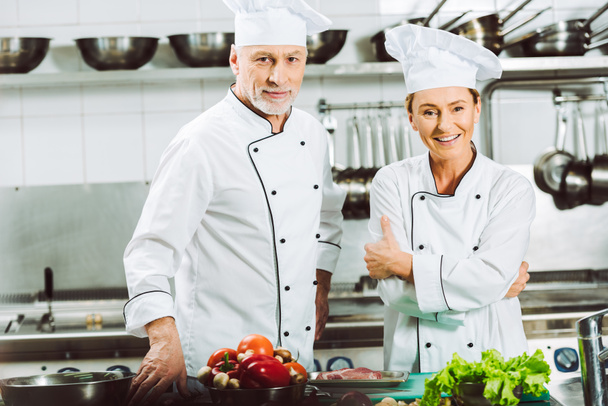 smiling female and male chefs in uniform and hats looking at camera while cooking in restaurant kitchen - Valokuva, kuva