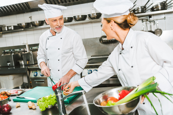 female and male chefs in uniform and hats looking at each other while cooking in restaurant kitchen - Foto, Bild