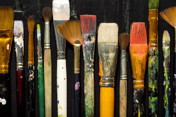 Artists tools, brushes in paint are in a row on a black wooden background. - 写真・画像
