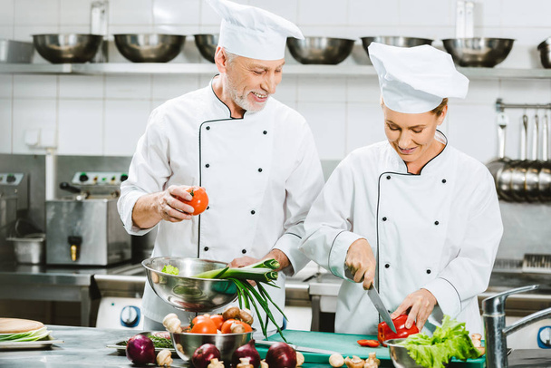 smiling female and male chefs in double-breasted jackets and hats cooking in restaurant kitchen - Fotoğraf, Görsel