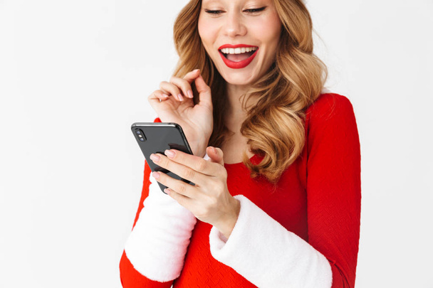 Portrait of happy woman 20s wearing Santa Claus red costume smiling and holding black smartphone isolated over white background - Foto, Imagen