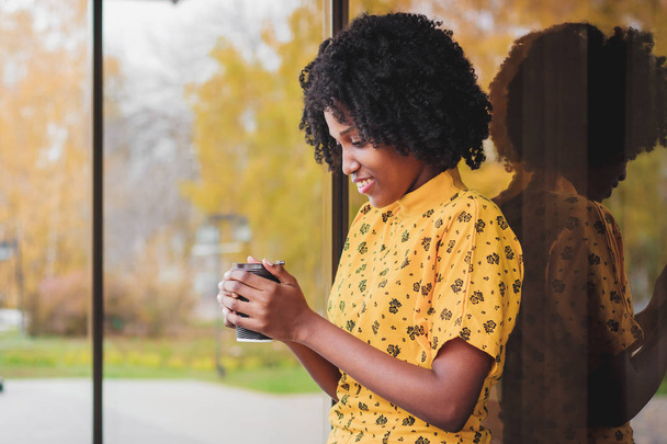 Mujer africana joven vestida con ropa casual amarilla sosteniendo una taza de bebida caliente, disfrutando del café
 - Foto, Imagen