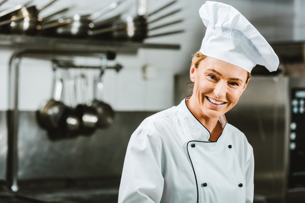 beautiful female chef in uniform and hat looking at camera in restaurant kitchen with copy space - Фото, изображение