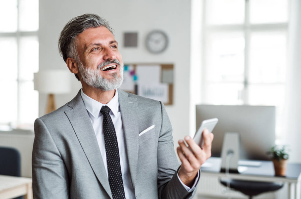 Cheerful mature businessman with smartphone standing in an office, laughing. - Photo, Image