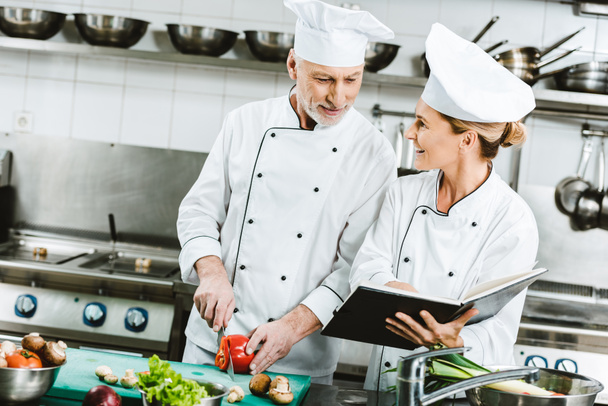 female and male chefs in uniform using recipe book during cooking in restaurant kitchen - Photo, Image