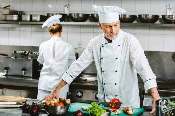 pensive male chef in double-breasted jacket during cooking in restaurant kitchen with female colleague on background - Photo, Image