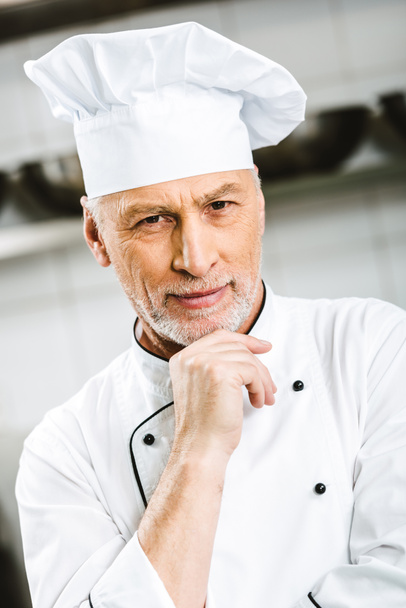 enfoque selectivo de guapo chef masculino apoyando la barbilla con la mano y mirando a la cámara en la cocina del restaurante
 - Foto, Imagen