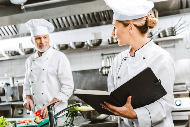 mujer chef en uniforme celebración de libro de recetas mientras colega de cocina en el fondo en la cocina del restaurante
 - Foto, Imagen