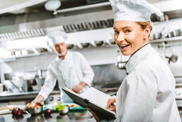 beautiful female chef in uniform holding recipe book and looking at camera in restaurant kitchen with colleague on background - Photo, Image