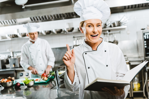 beautiful female chef in uniform holding recipe book and doing idea gesture in restaurant kitchen with colleague on background - Photo, Image